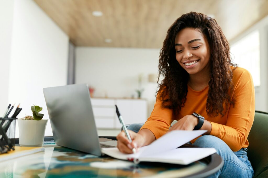 Black woman sitting at desk, using computer writing in notebook